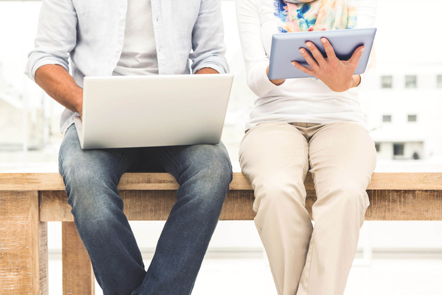 A man and woman sitting on a bench together using a laptop and tablet